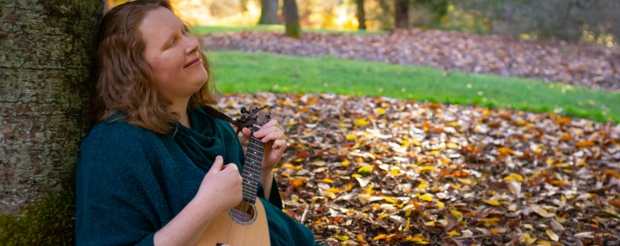 Redheaded woman playing the ukulele, sitting against a tree